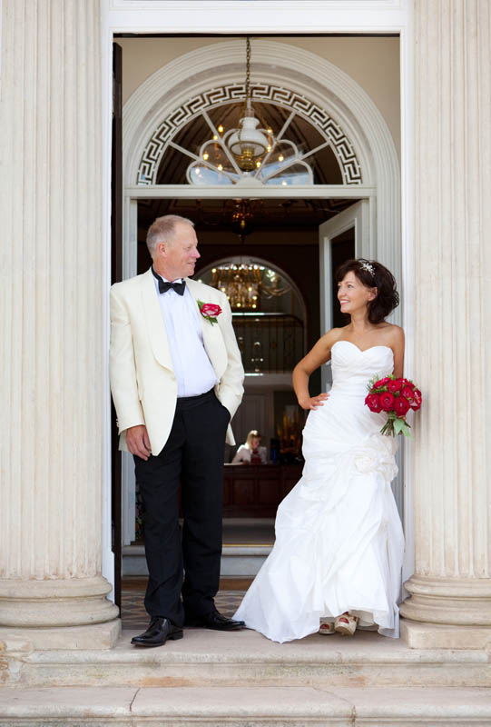 A bride and groom both in white lean against pillars on their wedding day at The Mount Somerset wedding venue
