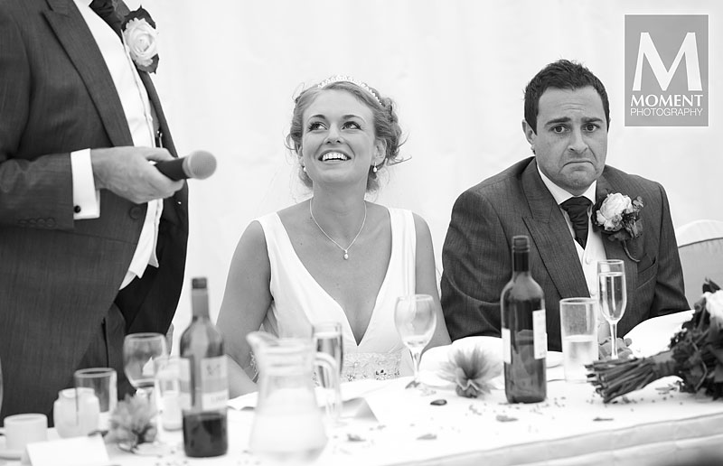 A black-and-white photo of a bride and groom listening to the speeches at the top table in the marquee at Lakeview Manor in Honiton