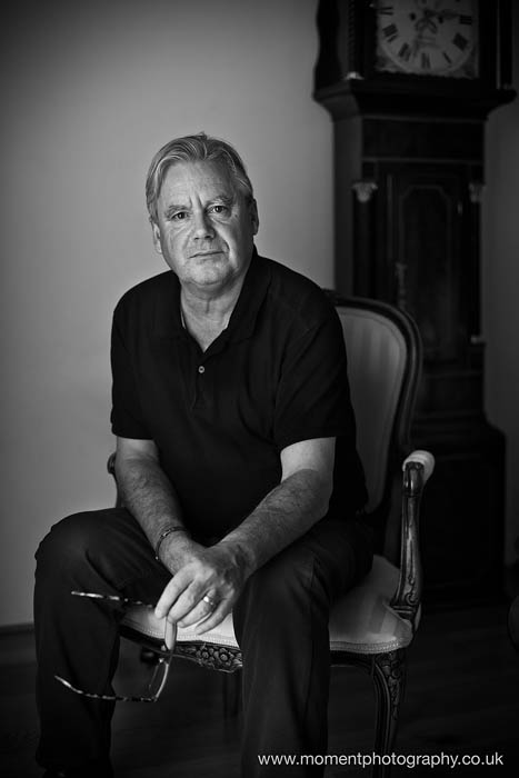 Black and white Portrait of author Neil Grimmett sitting in a chair holding his glasses.