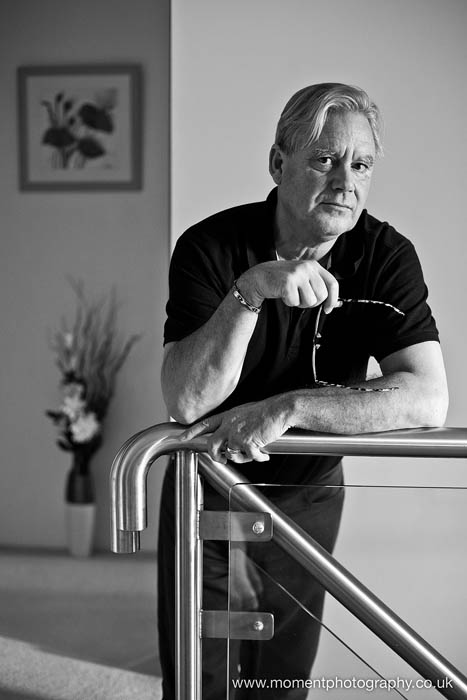 Black and white Portrait of author Neil Grimmett leaning on a stair rail holding his glasses.