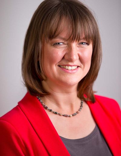 Staff business headshot of a brunette woman with a red jacket and grey top