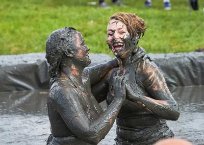 Two mud wrestling women covered in mud and slapping hands together with a smile on their faces